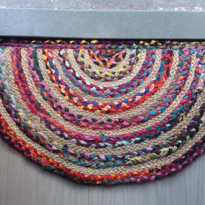 Könnte beinhalten: Ein halbkreisförmiger geflochtener Jute-Teppich mit einer lebendigen Farbmischung. Der Teppich weist konzentrische Bögen aus Rot, Rosa, Blau, Gelb und Grün auf und schafft einen strukturierten, dekorativen Bodenbelag. Geeignet für einen Eingangsbereich oder ähnliches.