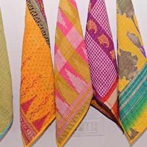 May include: Five colorful, patterned fabric pieces hanging against a white wall. The fabrics feature various designs, including geometric patterns, floral motifs, and patchwork. Colors include yellow, pink, purple, green, and red. The edges are trimmed with contrasting borders.