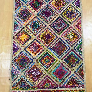 Puede incluir: Una alfombra rectangular tejida a mano con un patrón geométrico de diamantes. La alfombra presenta un borde beige y un diseño central de cuadrados trenzados coloridos en tonos de morado, azul, amarillo y rojo. La alfombra está hecha de fibras naturales.