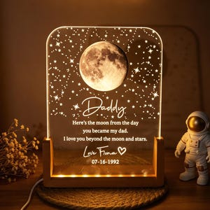 May include: A rectangular acrylic night light with a wooden base, featuring an engraved moon and stars design. The text reads: Daddy, Here's the moon from the day you became my dad. I love you beyond the moon and stars. Love Fiona 07-16-1992.