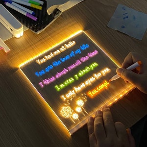 May include: A clear acrylic light-up sign with a message written in colourful markers. The message reads: "You had me at hello, You are the love of my life, I think about you all the time, I'm crazy about you, I only have eyes for you, You compl". The sign is illuminated with warm white LED lights around the perimeter.