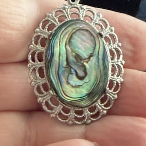 May include: Oval pendant with a silver filigree frame. The pendant features an iridescent abalone shell center with shades of green, blue, and purple. The pendant has a small loop at the top for a chain.