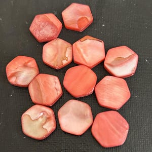 May include: A collection of eleven hexagonal, coral-colored beads. The beads have a pearlescent sheen and varying patterns of light and dark coral. The beads are arranged on a dark surface.