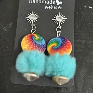 May include: Handmade earrings with a tie-dye design, turquoise fur, and silver accents. The earrings have a sunburst design at the top and a bell-shaped bottom. The text "Handmade with love" is printed on the black card.