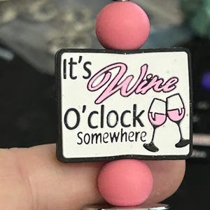 May include: A wine-themed charm with a white rectangular base featuring the text "It's Wine O'clock somewhere" in black and pink. Two wine glasses are depicted. The charm is accented with pink and silver beads.