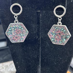 May include: A pair of silver-toned earrings. Each earring features a hexagonal pendant with a colorful, abstract design in shades of pink, teal, and purple. The pendants are suspended from a small silver ring at the top.