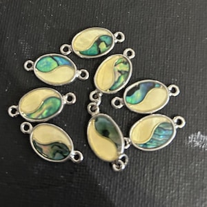 May include: Oval-shaped jewelry components with a silver-colored border. Each component features a cream-colored background with a swirling, iridescent green and blue design. The components have small loops on each end for attachment.