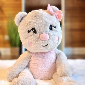 May include: A plush gray teddy bear with pink accents. The bear has a pink bow on its head, pink cheeks, and a pink nose. The bear is sitting on a white fluffy surface.