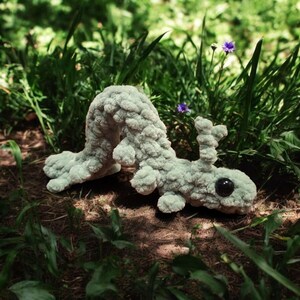 Pode incluir: Uma criatura de pelúcia de crochê verde claro com um corpo segmentado, pernas pequenas e um olho grande e preto. O brinquedo feito à mão está em uma cama de folhas marrons e grama verde, com algumas flores roxas ao fundo.