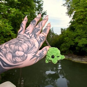 May include: A green crocheted keychain shaped like a small monster with black eyes. The keychain is hanging from a silver chain and is being held by a hand with a tattoo.