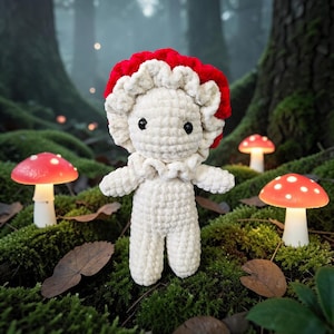 Puede incluir: Una figura de hongo de peluche hecha a mano con una gorra roja y blanca, cuerpo blanco y ojos negros. La figura está rodeada de luces de hongo brillantes y musgo en un entorno forestal. El peluche mide aproximadamente 15 cm de altura.
