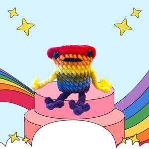 May include: A rainbow crocheted plush toy with a friendly face sits on a pink platform with a rainbow background. The toy has a red head, orange body, yellow arms, green legs, and purple feet.