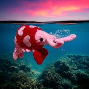 May include: A pink and red crocheted plush toy shaped like a sea creature with a long snout and a heart-shaped pattern. The toy is submerged in water with bubbles around it.