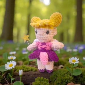 Könnte beinhalten: Eine handgefertigte Häkelfee-Puppe mit gelber Krone und sternförmigem Zauberstab. Die Puppe hat ein rosa-violettes Kleid und weiße Flügel. Die Puppe steht in einer natürlichen Umgebung mit Blumen und Grün.