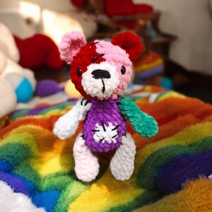 Puede incluir: Un oso de peluche de crochet con un esquema de color rosa, rojo, blanco y verde. El oso lleva un suéter morado con un diseño de copo de nieve blanco. El oso está sentado en una manta de colores arcoíris.