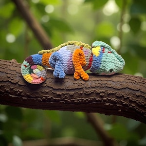 Op de afbeelding: Een kleurrijke, gehaakte kameleon rust op een tak. Het handgemaakte reptiel heeft een spiraalvormige staart en een kop met gele en blauwe accenten. Het lichaam is een mix van blauw, oranje, paars en groen garen, wat een levendige uitstraling geeft.