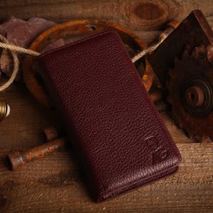 May include: A rectangular, deep burgundy wallet with a pebbled texture and a debossed logo in the bottom right corner. The wallet features a slim, folded design with visible stitching along the edges, showcasing a consistent, dark red leather-like finish.