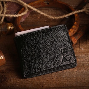 May include: Black leather wallet with a textured surface, featuring an embossed design. The wallet is open, revealing a white card. The background includes rustic elements on a wooden surface.