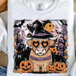 Op de afbeelding: Lichtgrijze sweatshirt met een Halloween-thema. Het ontwerp toont een Schotse hooglander met een heksenhoed en hartvormige zonnebril, omringd door pompoenen, geesten, vleermuizen en snoep. Het sweatshirt is van het merk Gildan.