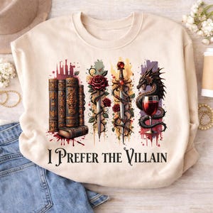 Puede incluir: Sudadera beige con un diseño gráfico que presenta libros apilados, espadas entrelazadas con rosas, un dragón y una copa de vino. El texto "I Prefer the Villain" está impreso debajo.