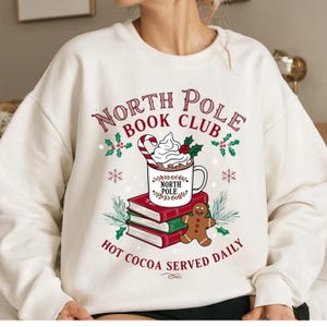 Op de afbeelding: Crèmekleurige sweatshirt met een feestelijk ontwerp. De afbeelding toont een mok warme chocolademelk met slagroom, een zuurstok en de tekst "North Pole Book Club". Onder de mok staan boeken en een peperkoekmannetje. De zin "Hot Cocoa Served Daily" is ook opgenomen.