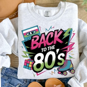 May include: A gray sweatshirt with a retro 80s design featuring a cassette tape, a Rubik's cube, roller skates, and the text "Back to the 80's".