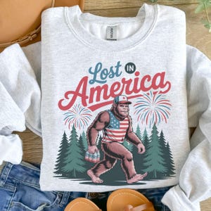 May include: A light grey sweatshirt with the text "Lost in America" in red and blue. The graphic shows a Bigfoot-like creature wearing an American flag vest and hat, carrying drinks, with fireworks and trees in the background.