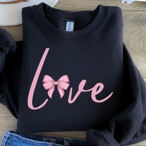 Op de afbeelding: Zwart sweatshirt met een roze strik en het woord "love" in een cursief schrift.