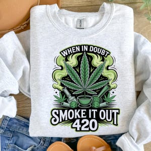 Pode incluir: Camisola cinza claro com um gráfico temático de cannabis. O gráfico inclui uma grande folha de cannabis verde, uma tigela e o texto "WHEN IN DOUBT SMOKE IT OUT 420" em preto e verde.