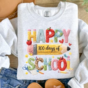 May include: Light gray sweatshirt with a colorful graphic that reads "HAPPY 100 days of SCHOOL." The design includes a large yellow pencil, red apples, stars, and other school-themed elements. The sweatshirt is made of a soft material.