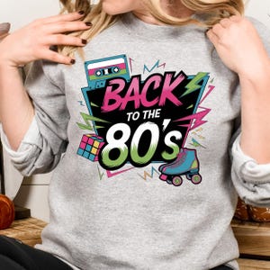 May include: A gray sweatshirt with a graphic design featuring a cassette tape, a Rubik's cube, roller skates, and the text "Back to the 80's".