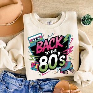 May include: A cream-colored sweatshirt with a graphic print of a cassette tape, a Rubik's cube, roller skates, and the text "Back to the 80's".