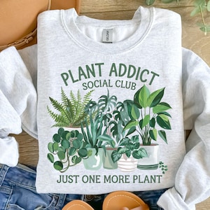 May include: A light gray sweatshirt with the text "Plant Addict Social Club" and a graphic of various potted plants. The text "Just One More Plant" is printed below the graphic.