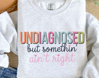 Pode incluir: Camisola cinzenta com o texto "UNDIAGNOSED but somethin' ain't right" em letras coloridas com corações.