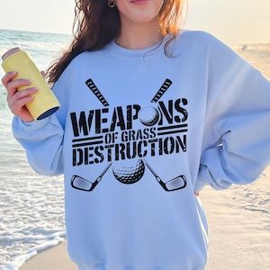 May include: Light blue sweatshirt with a black graphic of two golf clubs crossed with a golf ball in the center. The text "WEAPONS OF GRASS DESTRUCTION" is printed in black above the graphic.