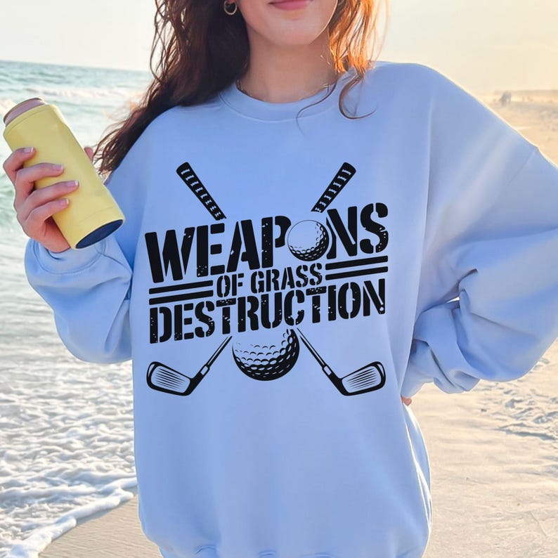 May include: Light blue sweatshirt with a black graphic of two golf clubs crossed with a golf ball in the center. The text "WEAPONS OF GRASS DESTRUCTION" is printed in black above the graphic.