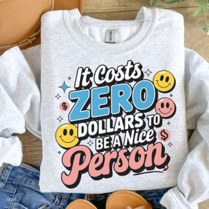 May include: A grey sweatshirt with the text "It Costs Zero Dollars To Be A Nice Person" in colourful, stylised lettering. The text is surrounded by smiley faces and dollar signs.