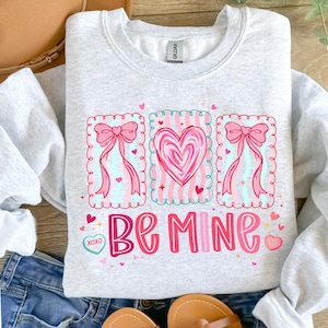 May include: A light gray sweatshirt with a Valentine's Day design. The design includes pink bows, hearts, and the words "Be Mine" in pink and red lettering. The sweatshirt is folded on a wooden surface.