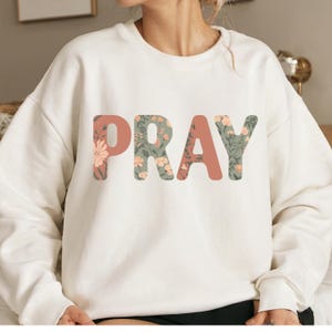 Op de afbeelding: Crèmekleurige sweatshirt met het woord "PRAY" in een bloemenontwerp. De letters zijn een mix van gedempt koraal en bloempatronen, waardoor een zachte, vrouwelijke esthetiek ontstaat. De sweatshirt is een casual, comfortabele stijl.
