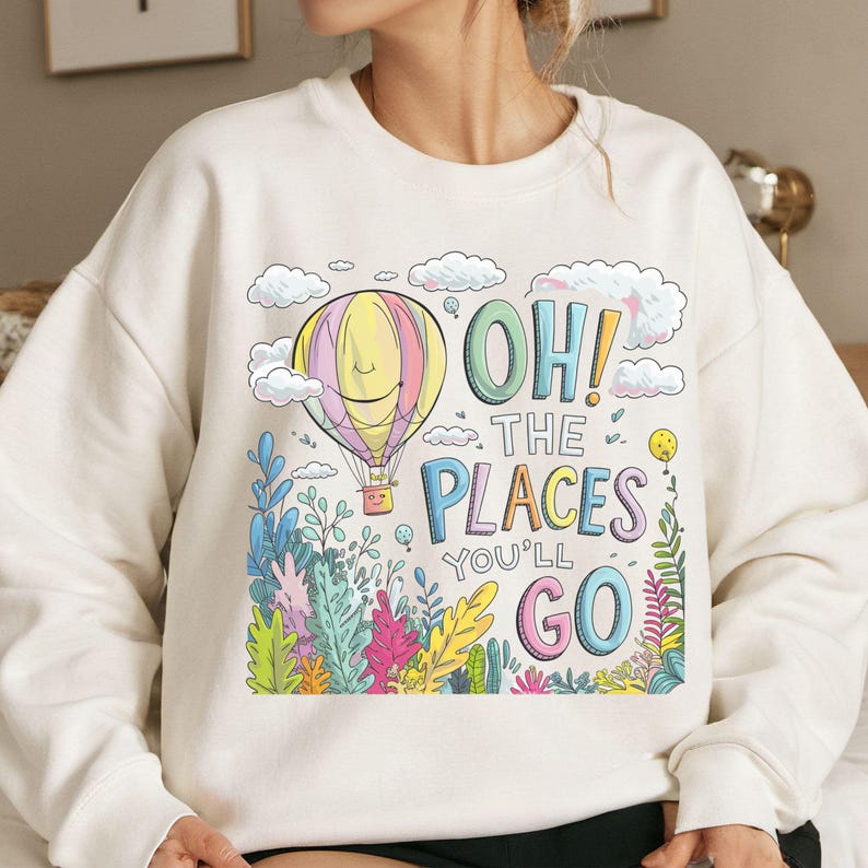 May include: White sweatshirt featuring a colourful hot air balloon, clouds and foliage graphic. The text "Oh! The Places You'll Go" is printed on the sweatshirt.