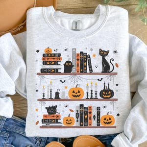 Op de afbeelding: Lichtgrijze sweatshirt met een Halloween-thema boekenplankontwerp. Het ontwerp bevat gestapelde boeken, pompoenen, een zwarte kat, kaarsen, spinnen en een spook. De sweatshirt heeft een ronde hals en lange mouwen.