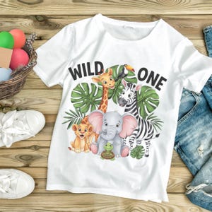 May include: White t-shirt with a graphic of a giraffe, zebra, elephant, lion, and turtle. The text "WILD ONE" is printed on the shirt.