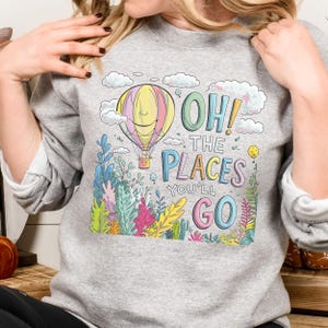 May include: A grey sweatshirt featuring a colourful illustration of a hot air balloon, clouds and foliage. The text "Oh! The Places You'll Go" is printed on the sweatshirt.
