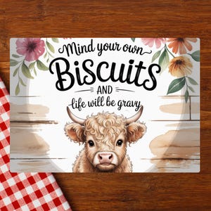 Può includere: Cartello rettangolare con la scritta "Mind your own Biscuits AND life will be gravy". Il cartello presenta un'illustrazione ad acquerello di una mucca e accenti floreali rosa e arancioni. Un panno a scacchi rosso e bianco si trova nell'angolo in basso a sinistra.