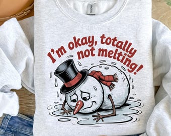 May include: Light gray sweatshirt with a graphic of a melting snowman wearing a top hat and scarf. The text "I'm okay, totally not melting!" is printed in red. A humorous winter-themed design.