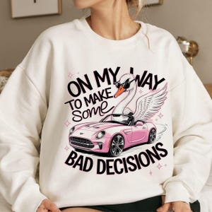 May include: White sweatshirt with a pink car and a swan wearing sunglasses. The text on the sweatshirt reads "On my way to make some bad decisions".
