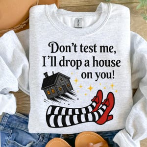 May include: Light gray sweatshirt with the text "Don't test me, I'll drop a house on you!". The graphic features a black house, striped legs, and red shoes, with yellow stars.