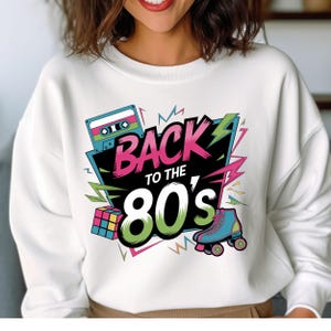 May include: White sweatshirt with a graphic print of a cassette tape, roller skates, and a Rubik's cube. The graphic says "Back to the 80's" in pink, green, and blue letters.
