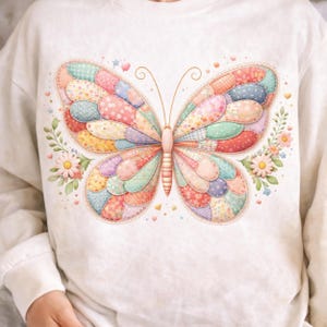 May include: A cream-colored sweatshirt featuring a large, colorful butterfly design. The butterfly has patchwork wings in various pastel colors, with floral and heart accents. The design is whimsical and detailed.