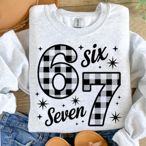May include: A light gray sweatshirt with the numbers "6" and "7" in a black and white buffalo plaid pattern. The word "six" and "Seven" are written in black script. Starburst graphics are also included.